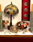 photo of small table with patterned, white tablecloth, two verdigris-color plaster candlesticks with lit orange candles, colorful glass lamp, decorative bowls with pine cones and a Venetian mask, pillow with moose image, and on off-white wall behind, a ta