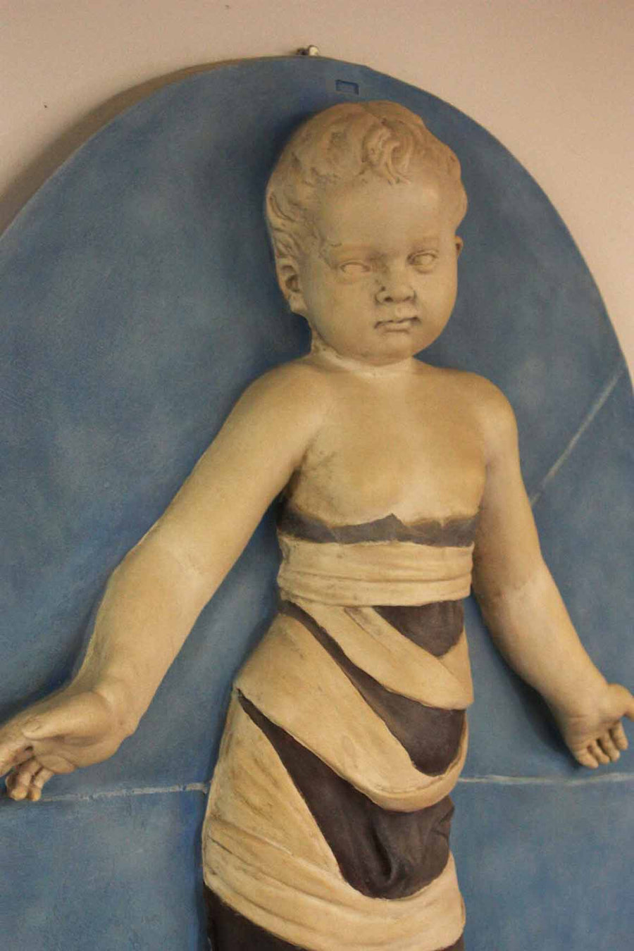 closeup photo of plaster cast relief of child with mummy-like wrappings on a blue oval background on a tan-colored wall