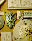 photo of plaster cast sculpture reliefs on orange-brown wall, including verdigris-colored relief of moustached man's face made of leaves