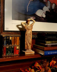 photo of plaster cast statue of a female in pink-orange tinted robes with her arms raised to her head placed on a wooden fireplace mantel beside stacks of books with a framed print on wall behind