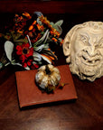 photo of plaster cast sculpture of grotesque faun face on a dark table next to fall flowers, a pumpkin, and a red book