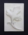 Photo of a plaster cast of a rectangle slab with a study of an acanthus leaf poited upward on the front, on a black background