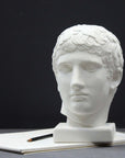 photo of plaster cast of sculpture of male head with flat, curly hair on top of sketchbook along with a pencil on dark gray surface