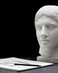 photo of plaster cast sculpture of head of the Doryphoros, male with curly hair, on dark gray surface with sketchbook and pencil beside it
