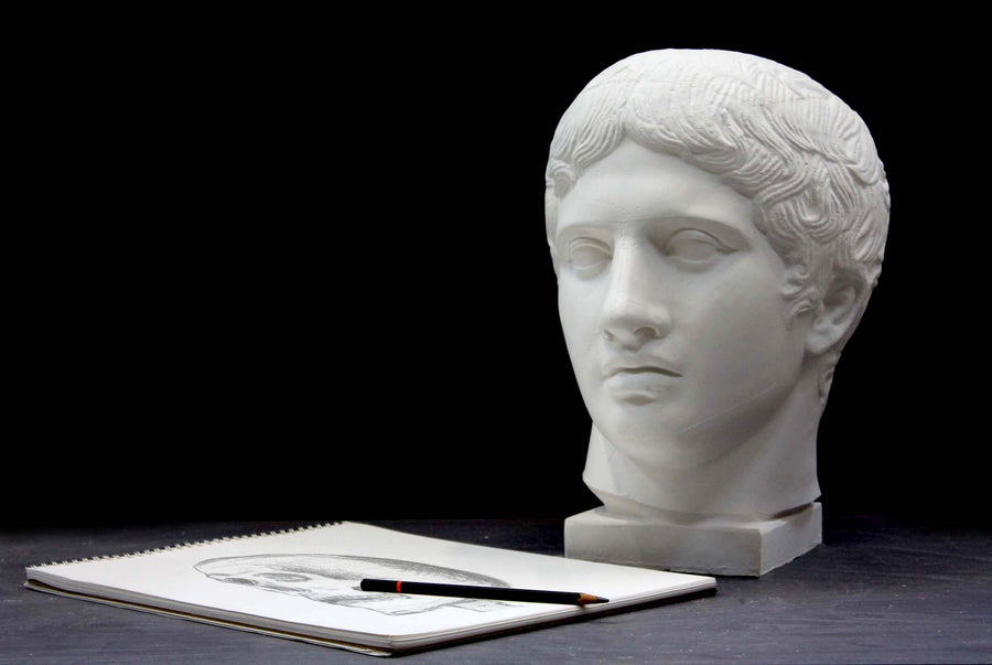 photo of plaster cast sculpture of head of the Doryphoros, male with curly hair, on dark gray surface with sketchbook and pencil beside it