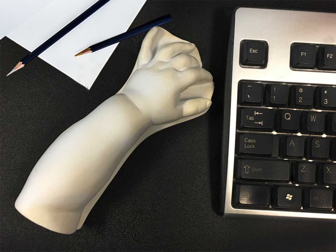 photo of white plaster cast sculpture of baby lower arm sitting on a desk between a keyboard on the right and a sheet of paper and two drawing pencils on the left