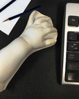 photo of white plaster cast sculpture of baby lower arm sitting on a desk between a keyboard on the right and a sheet of paper and two drawing pencils on the left