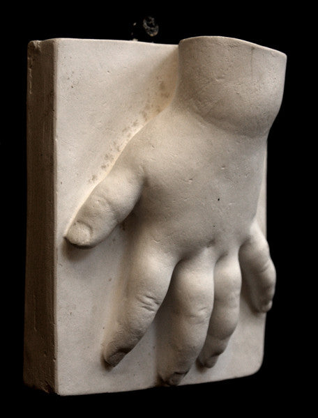 photo of off-white plaster cast sculpture of baby hand on panel against black background