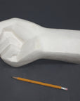 photo of white plaster cast sculpture of hand in fist in block forms with yellow pencil beside it against gray background