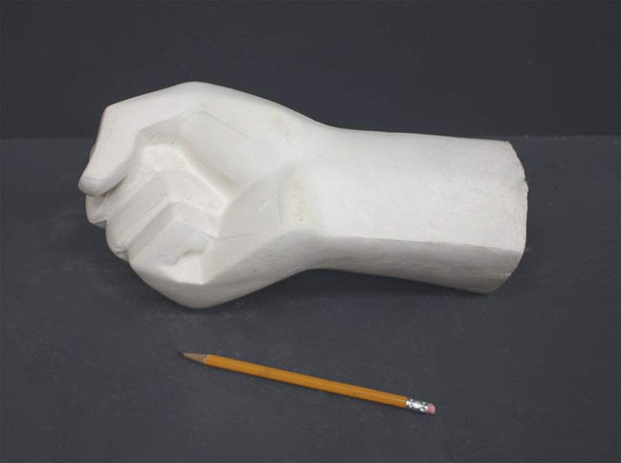 photo of white plaster cast sculpture of hand in fist in block forms with yellow pencil beside it against gray background