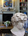 Photo of a colorful living room with blue striped sofas, fireplace, and bookshelves and a plaster cast sculpture of male head of Antinous with crown of leaves in the foreground