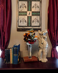 photo of plaster cast sculpture of ornamental seated winged lion on a dark sofa table with antique books and flowers, red curtains behind, and painting on wall
