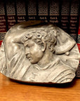 photo of plaster cast sculpture relief of upper body of male with arm raised behind head and holding a hammer leaning against an encyclopedia series in a wooden bookshelf