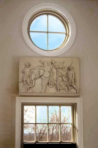 Photo of a plaster cast of figures and an element on a tan wall above a door and below a round window with blue sky coming through