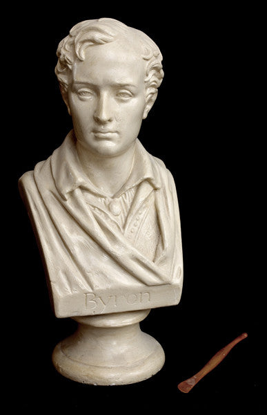 photo of white plaster cast sculpture bust of man, namely the poet Lord Byron, on socle base and sculpting tool beside it on black background