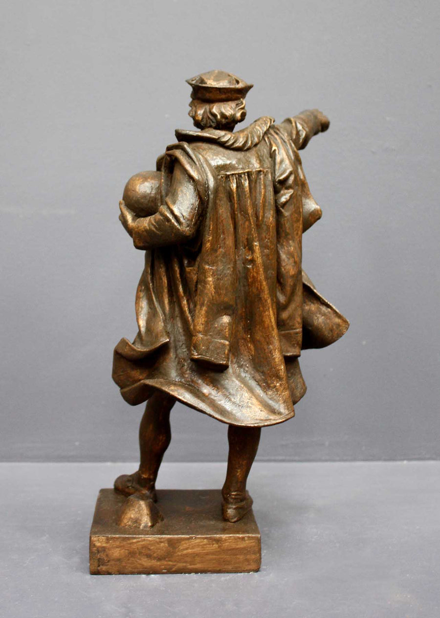 photo of bronze-colored plaster cast sculpture of standing man, namely Christopher Columbus, in robes and hat, pointing with right hand and holding a globe in his left arm against gray background