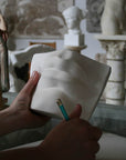 Photo of a woman's left hand holding the plaster cast of David's mouth by Michelangelo and right hand holding a drawing pencil with sculptures and a sketchpad in the background