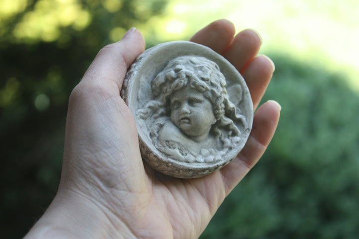 Photo of small round plaster sculpture medallion of head of an infant held by a light-skinned female hand with a blurry green background