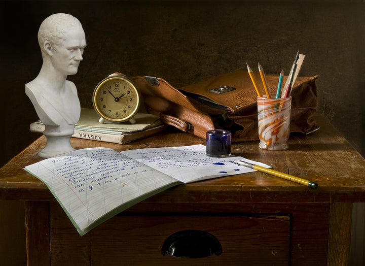 photo of dark wooden desk with open notebook and pen and ink, jar of pencils, small clock, leather bag, books, and plaster cast bust of Alexander Hamilton