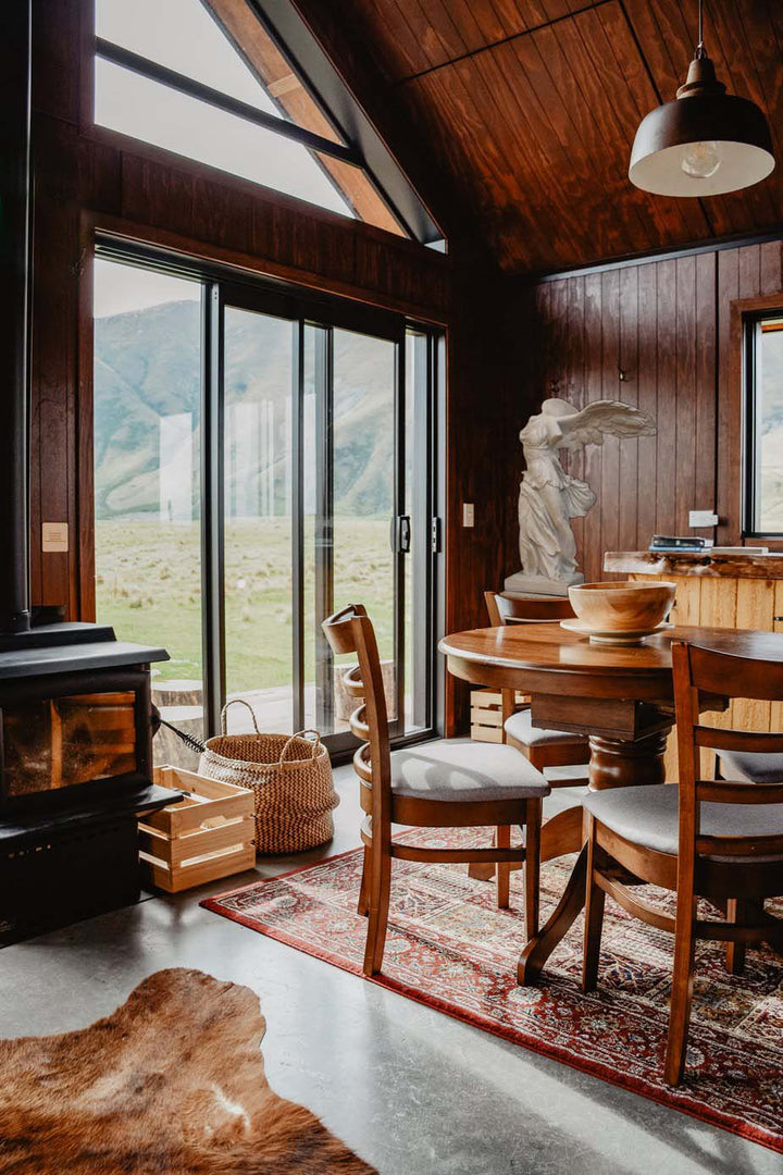photo of cabin with glass-door wall, table and chairs with hanging lamp above, wood stove, and Victory of Samothrace plaster cast sculpture in the corner on a table