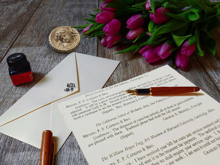 photo closeup of gray textured desktop with written letter, envelope, wooden-colored pen and pen cap, small square jar of ink, off-white small round plaster relief of Infant Bacchus, and top of bouquet of purple tulips lying on its side