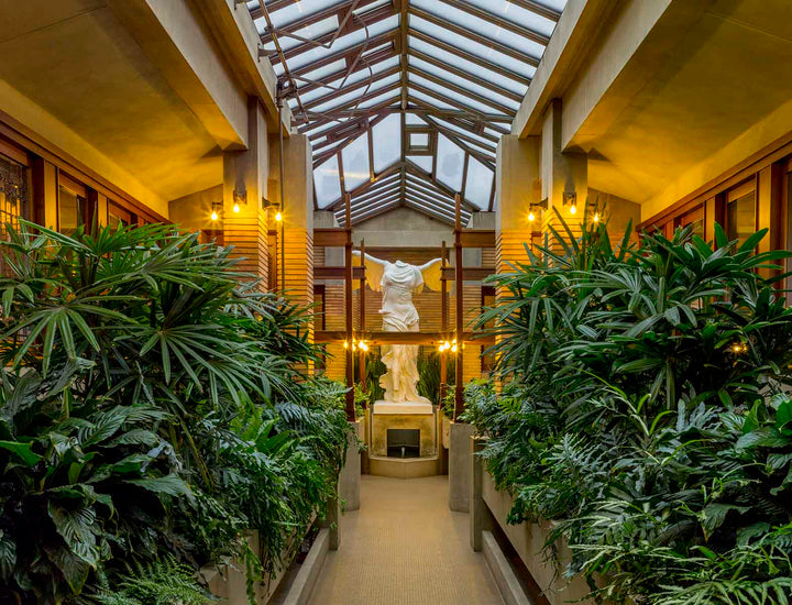photo of Victory of Samothrace sculpture at the end of a path bordered by green plants in a glass-roofed conservatory with tan colors and warm lighting