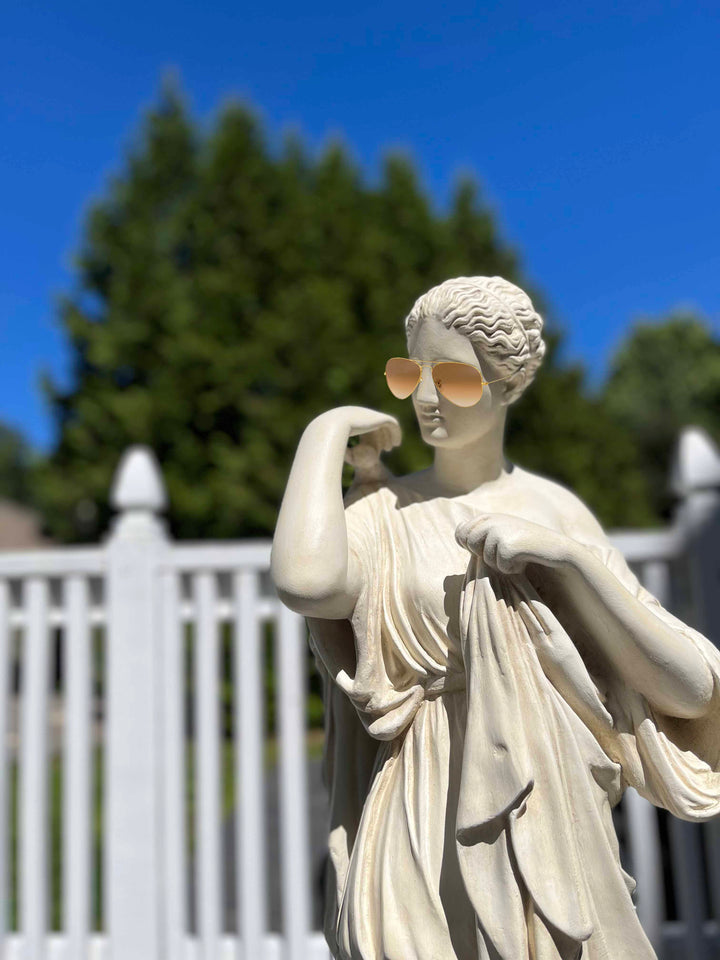 photo of white plaster cast sculpture of female figure in drapery with white fence, trees, and blue sky in background