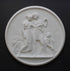 photo of white plaster cast relief sculpture of male and female figures walking through field of wheat and a female figure kneeling in the field cutting wheat against dark gray background