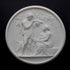 photo of white plaster cast relief sculpture of female figure with baby on lap, a standing male figure, and a dog against dark gray background