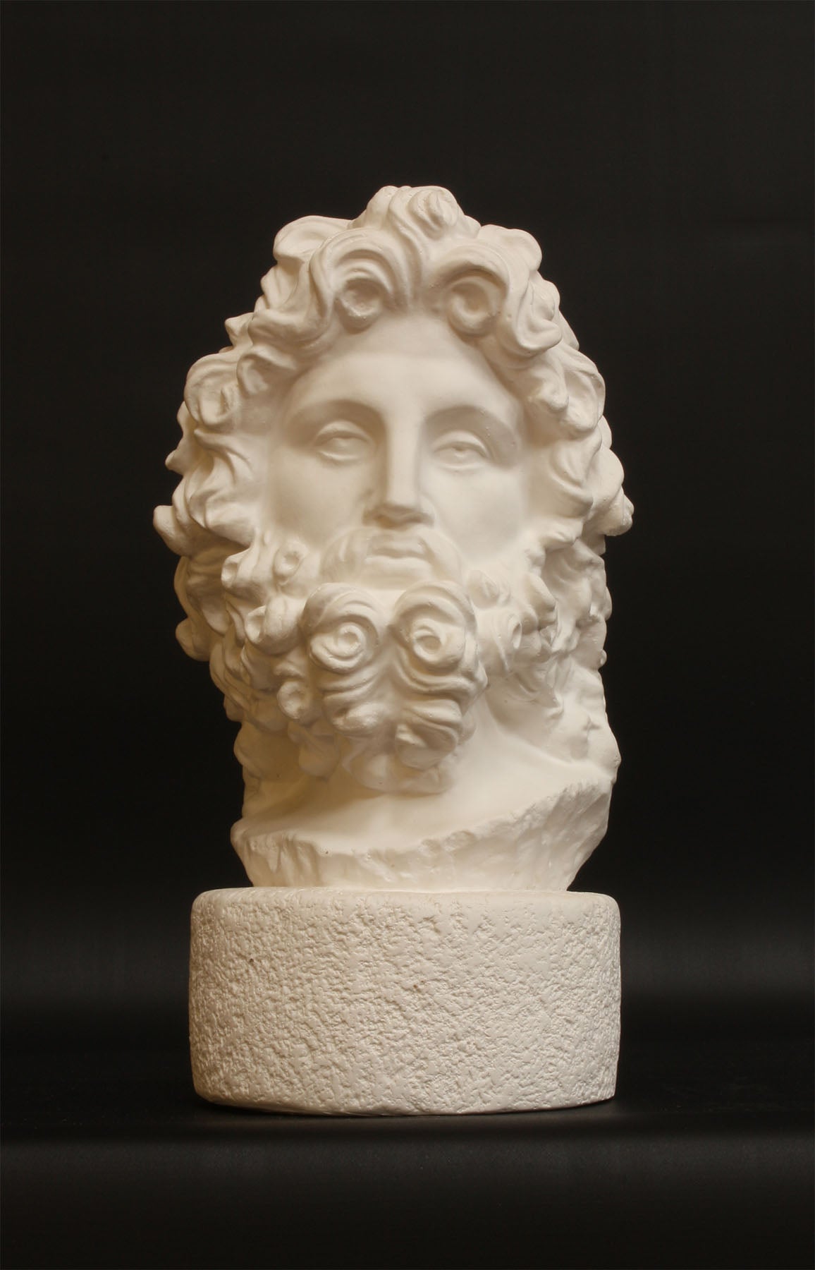 Photo with black background of plaster cast sculpture of male bust, namely the god Zeus, with curly hair and beard