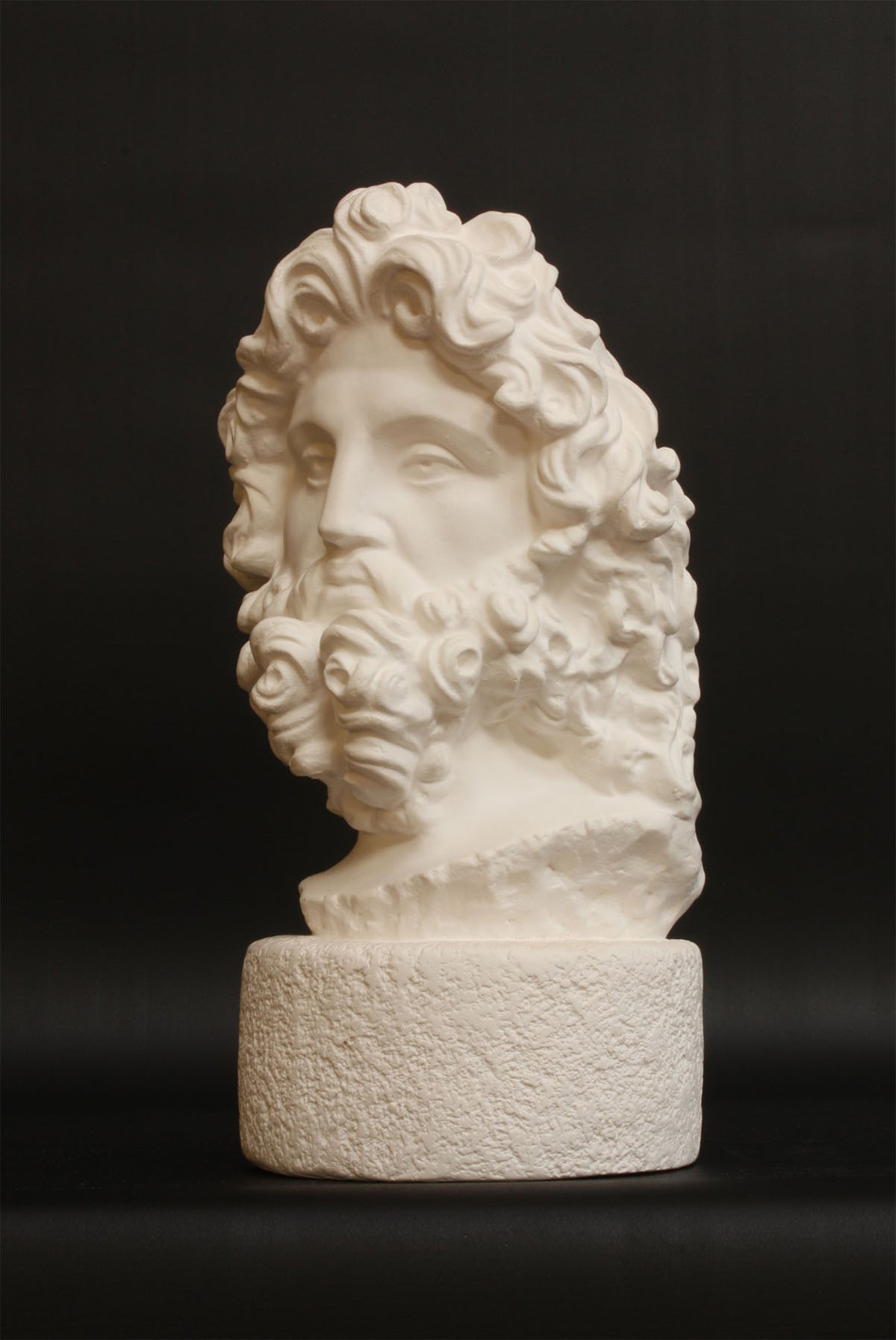 Photo with black background of plaster cast sculpture of male bust, namely the god Zeus, with curly hair and beard
