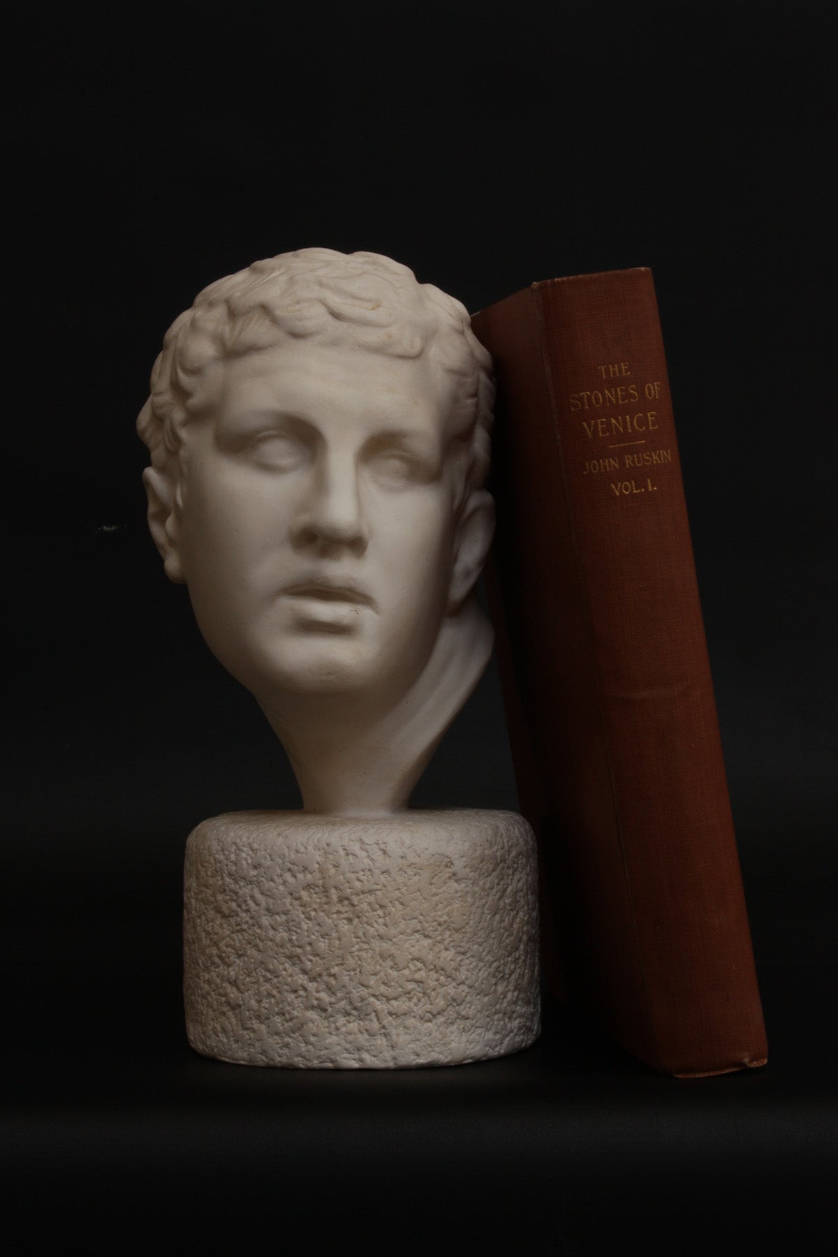 photo of white plaster cast reproduction of Ancient Greek bust of male from Borghese Warrior statue with red book leaning against it on black background