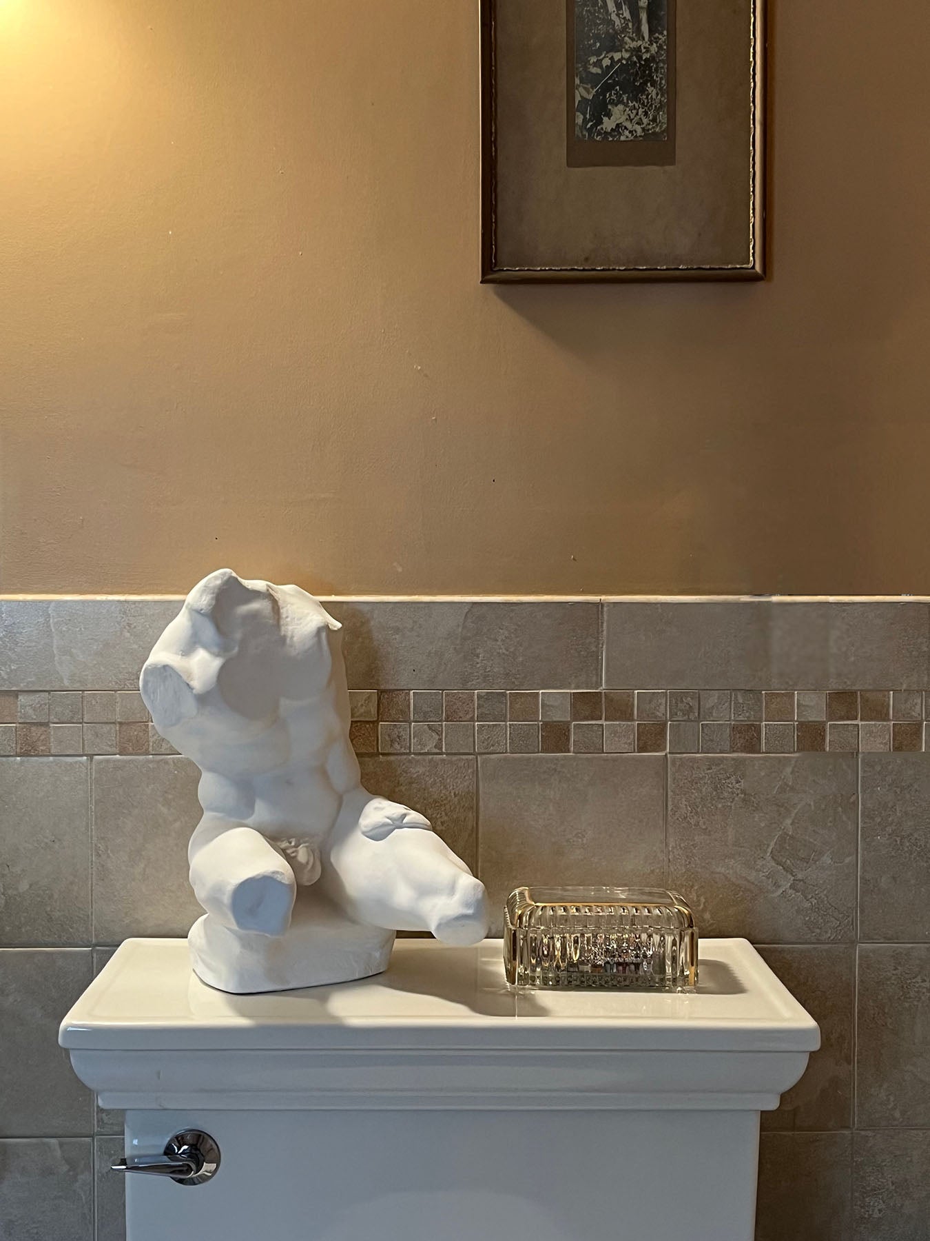 image of white plaster sculpture of nude male torso atop white toilet with gray and orange-brown tiling and orange-brown wall behind