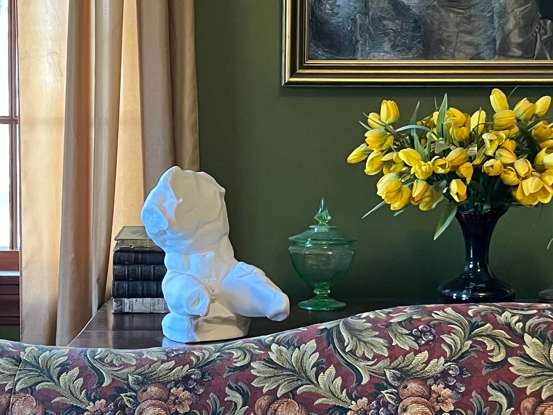 image of white plaster sculpture of nude male torso on brown sofa table behind red and green floral-patterned sofa next to antique books and black vase with yellow tulips with green wall and tan curtains behind