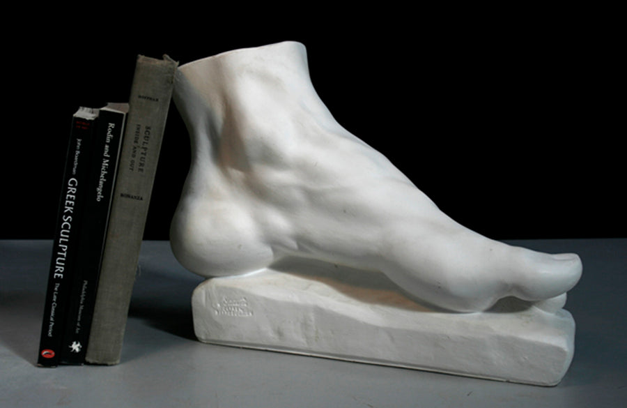 photo of white plaster cast sculpture of left foot on base with green and black books leaning against it against gray and black background