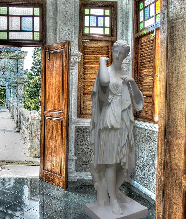 photo of white plaster cast of female figure, namely the goddess Diana, in robes about to clasp two pieces together at proper right shoulder, in corner of room with green marble floor, wooden doors and window treatments, and wall ornamentation, door open to outside stone terrace