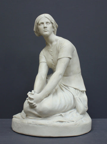 photo of white plaster cast sculpture of female, namely Joan of Arc, kneeling with hands on lap atop round pedestal on gray background