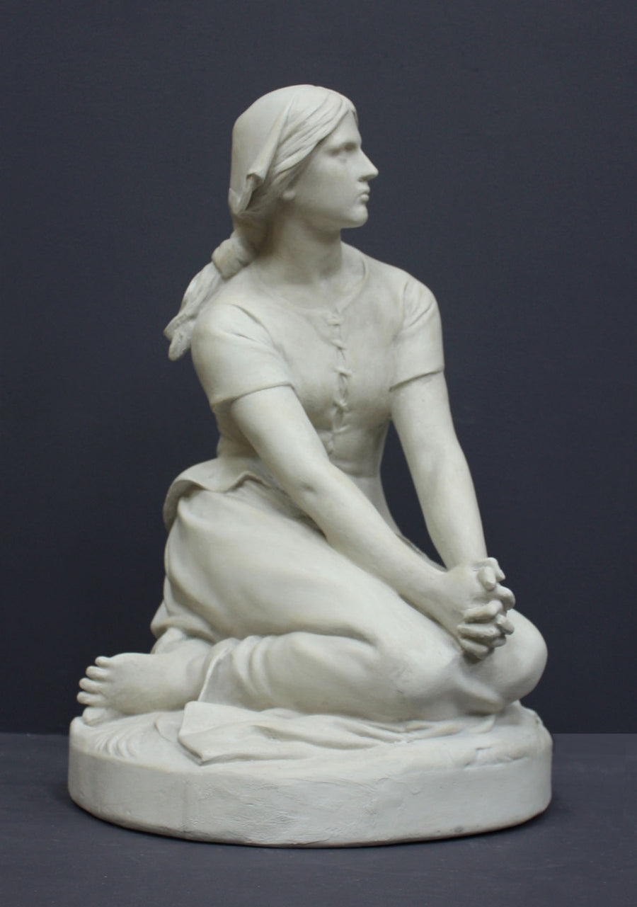 photo of white plaster cast sculpture of female, namely Joan of Arc, kneeling with hands on lap atop round pedestal on gray background