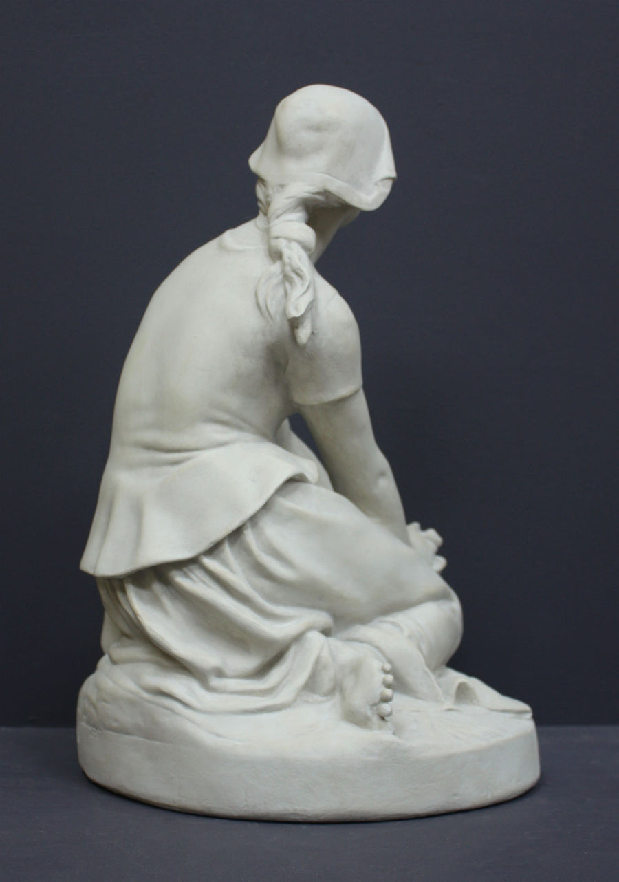photo of back view of white plaster cast sculpture of female, namely Joan of Arc, kneeling with hands on lap atop round pedestal on gray background