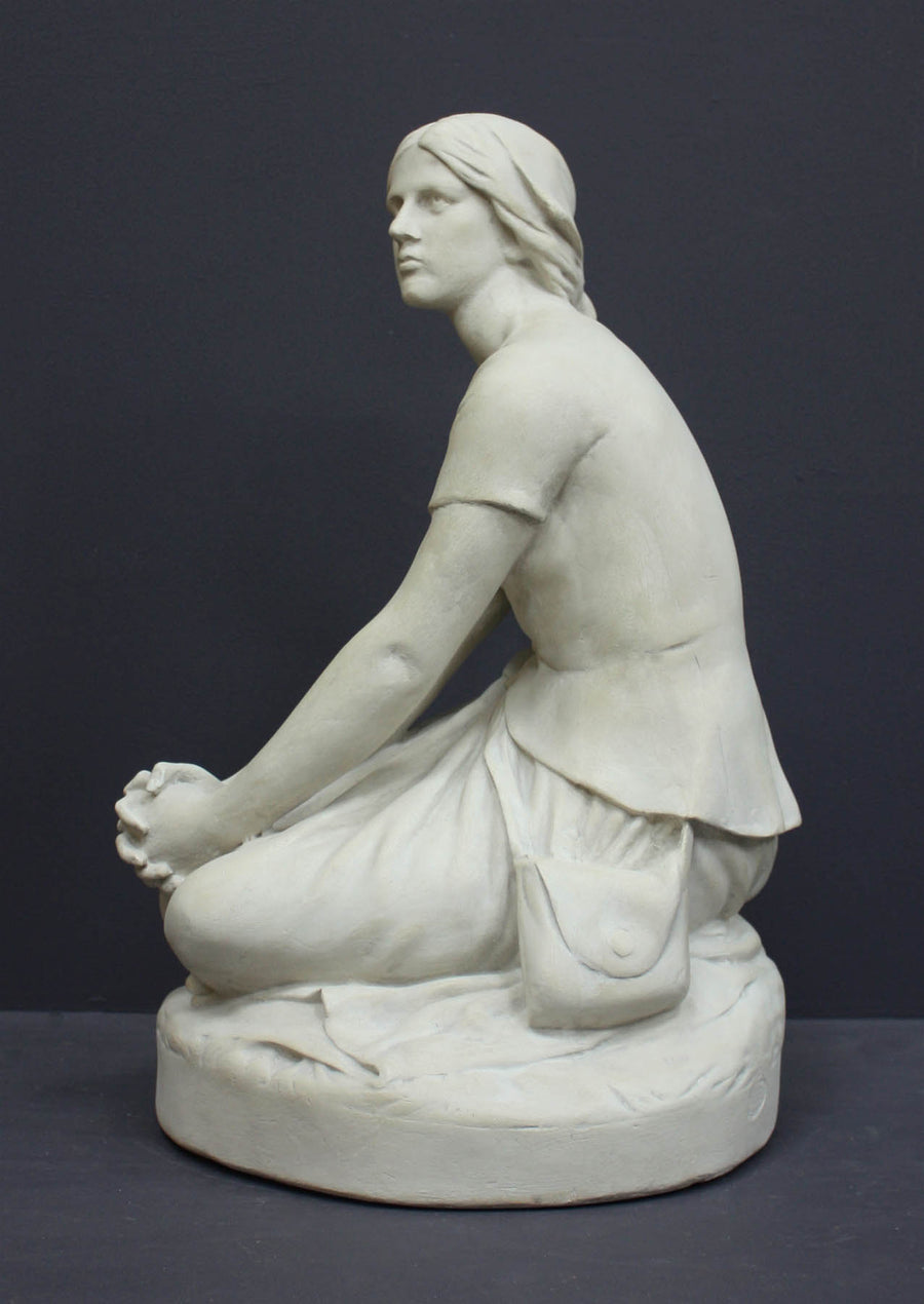 photo of white plaster cast sculpture of female, namely Joan of Arc, kneeling with hands on lap atop round pedestal on gray background