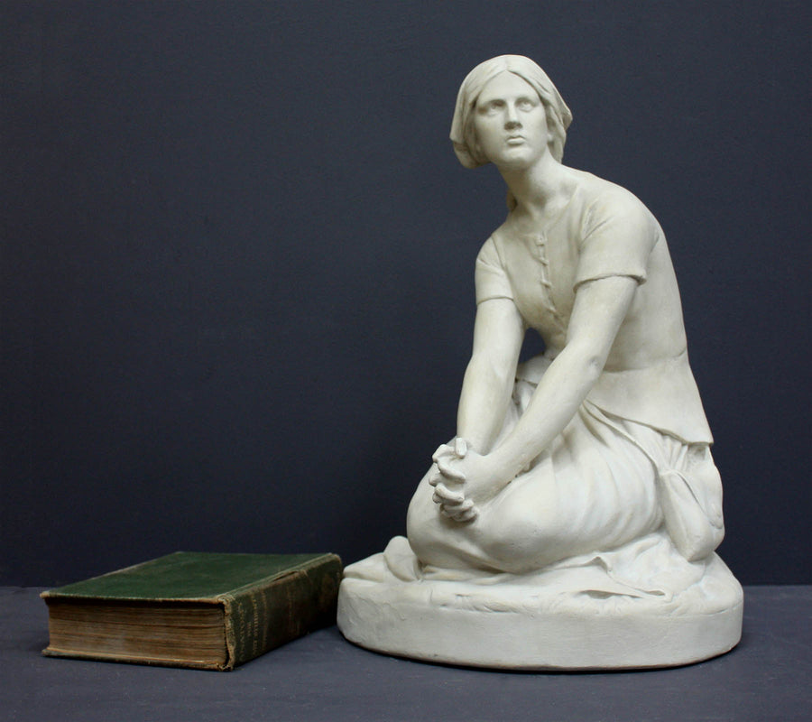 photo of white plaster cast sculpture of female, namely Joan of Arc, kneeling with hands on lap atop round pedestal with green book laid beside it on gray background