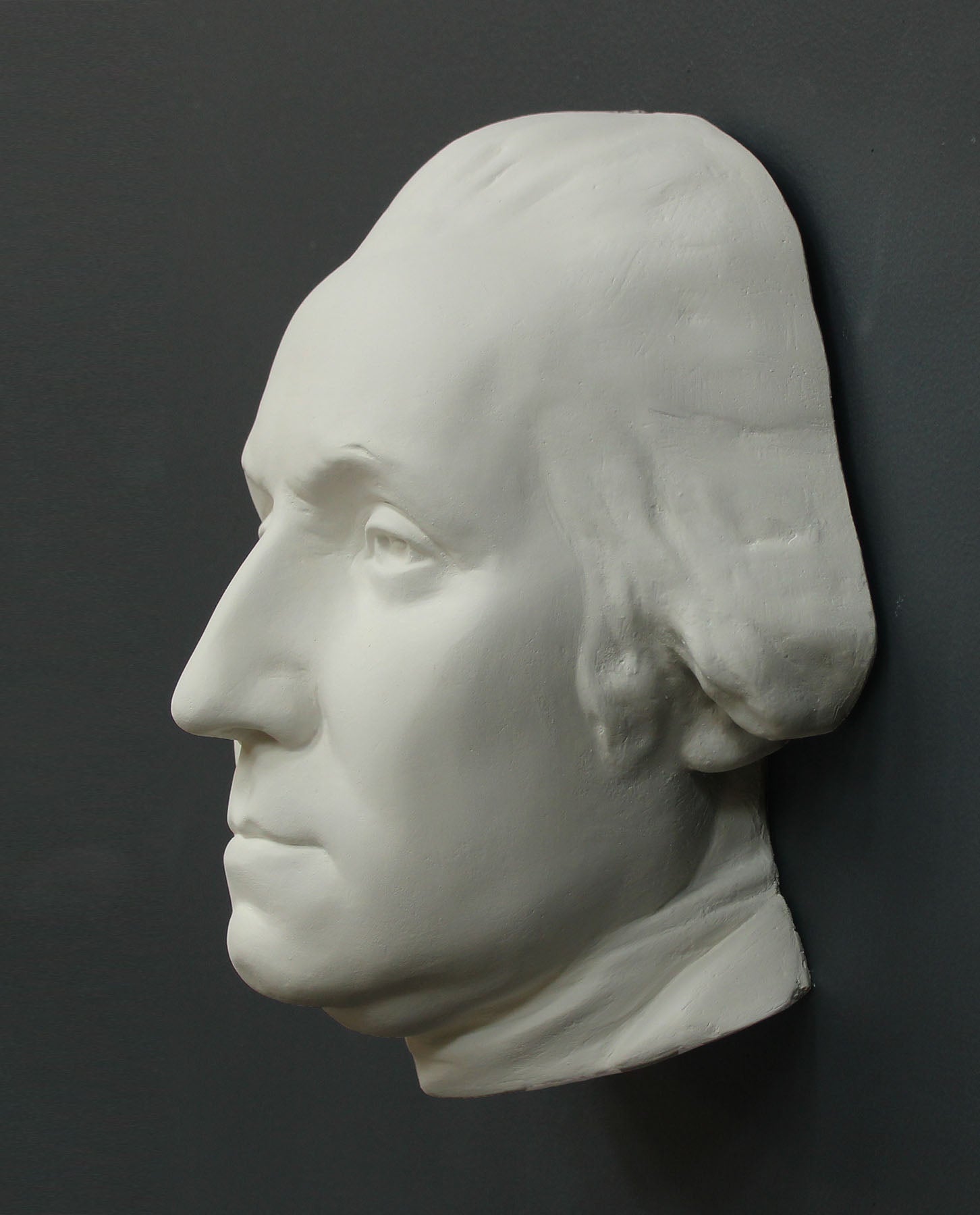 Photo of white plaster cast sculpture of mask/face of male in Revolutionary garb, namely George Washington, on gray background