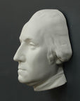 Photo of white plaster cast sculpture of mask/face of male in Revolutionary garb, namely George Washington, on gray background
