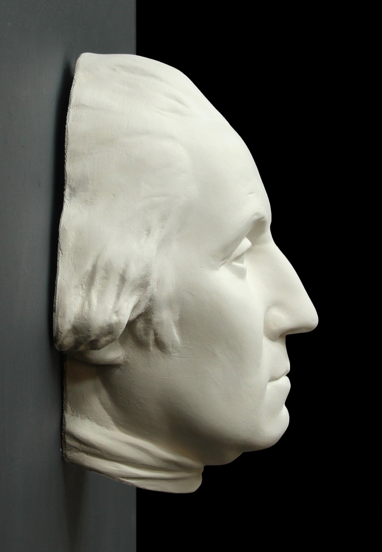 profile photo of white plaster cast sculpture of mask/face of male in Revolutionary garb, namely George Washington, on black and gray background
