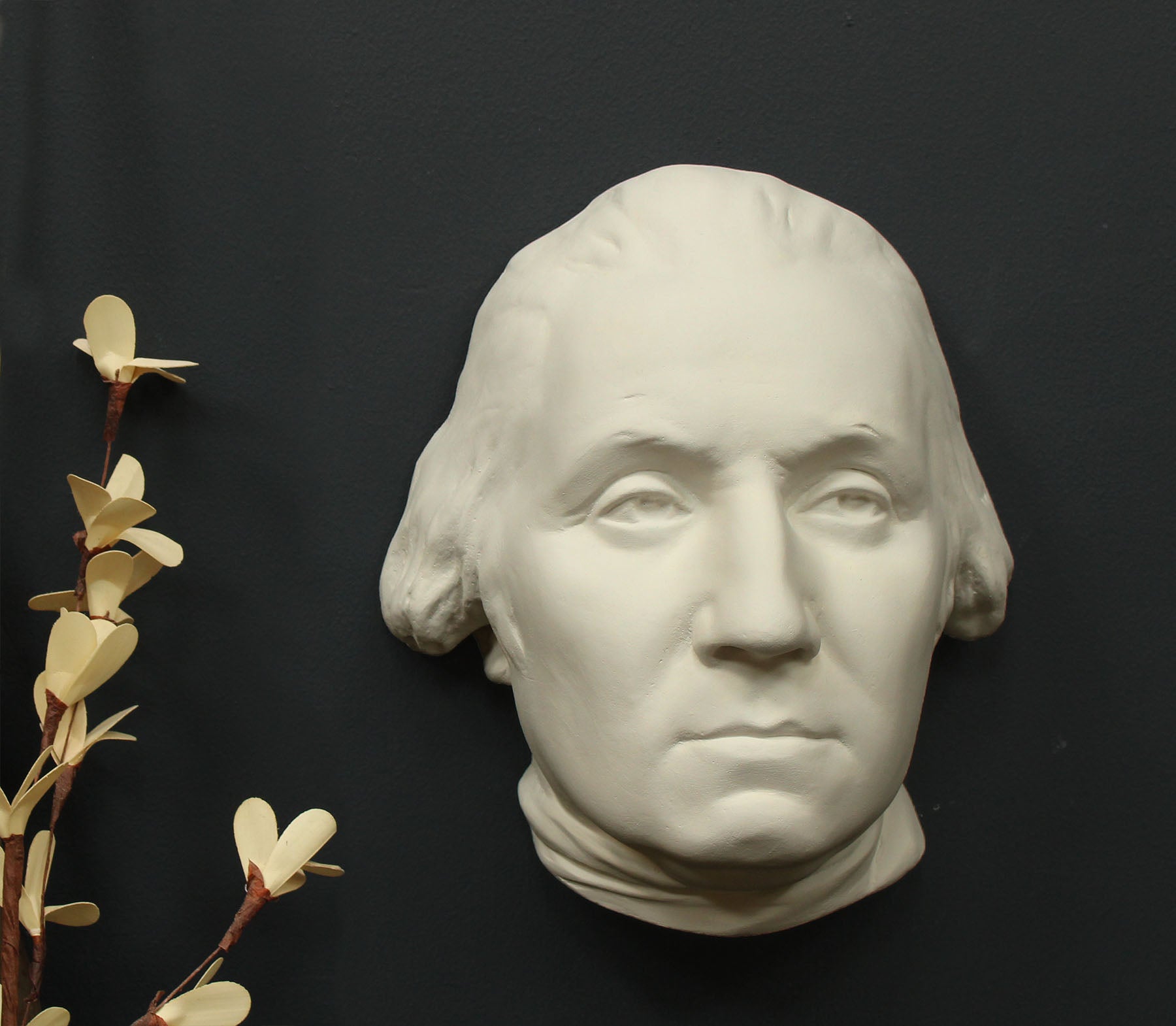 Photo of white plaster cast sculpture of mask/face of male in Revolutionary garb, namely George Washington, with ivory flowers to its right on gray background