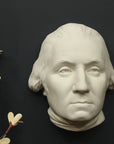Photo of white plaster cast sculpture of mask/face of male in Revolutionary garb, namely George Washington, with ivory flowers to its right on gray background