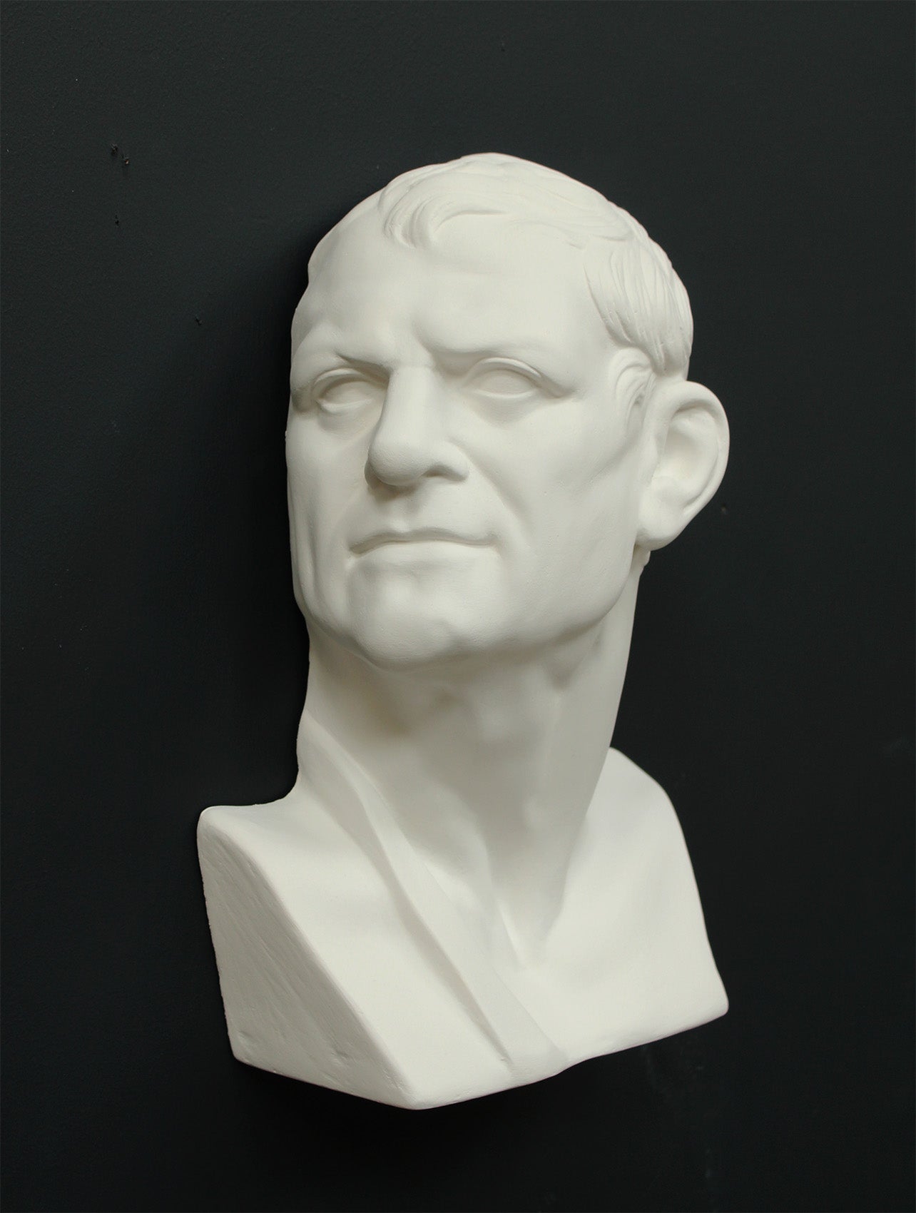 Photo of white plaster cast sculpture of mask/face of male facing right on gray background