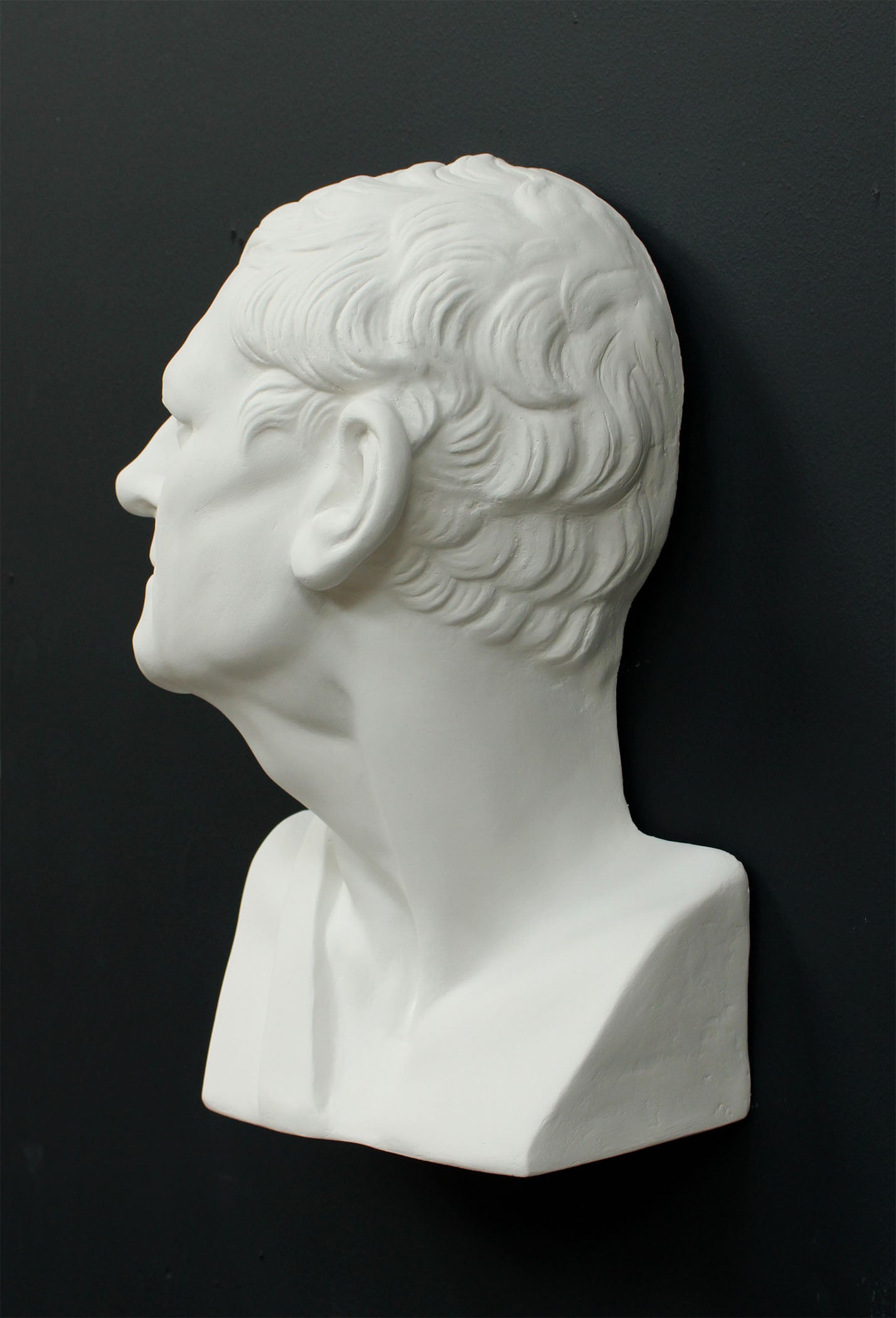Photo of white plaster cast sculpture of mask/face of male facing right on gray background