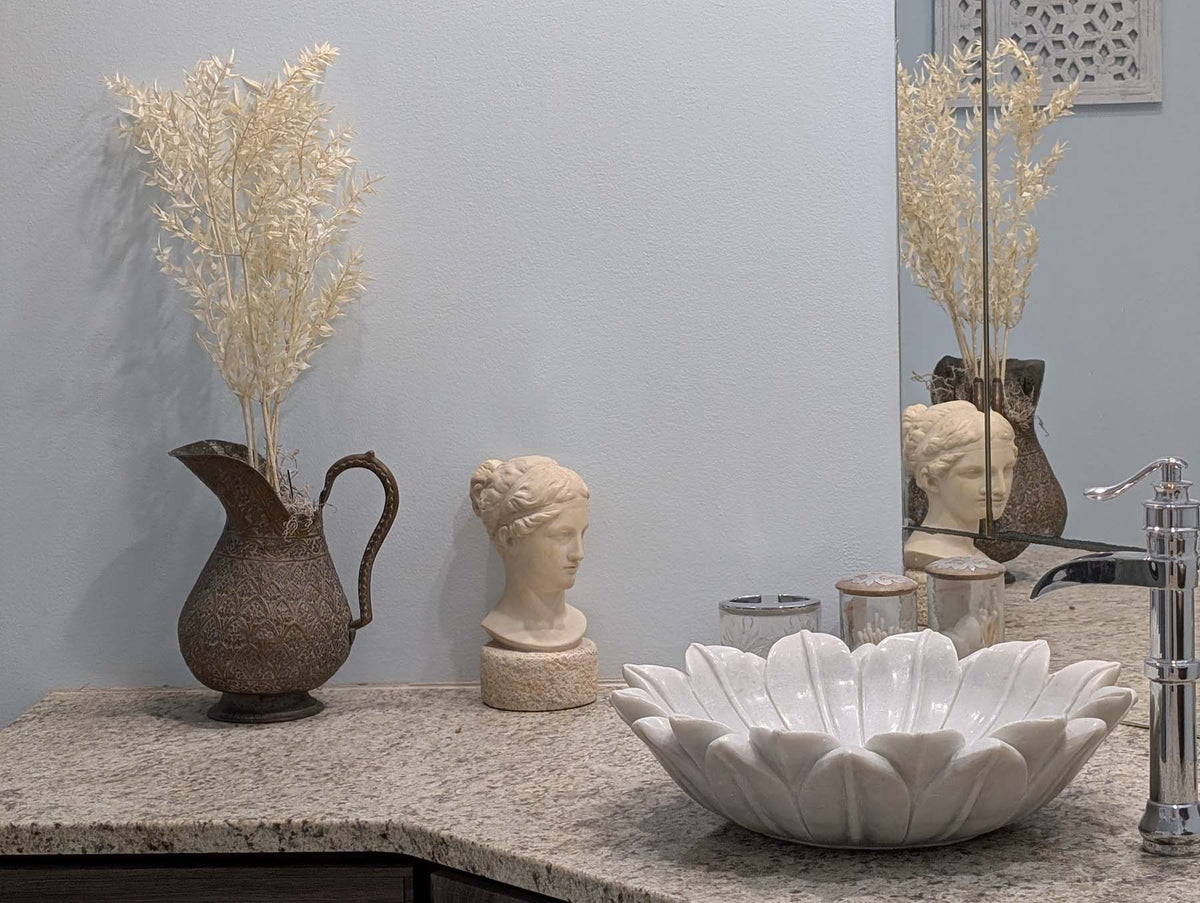 Photo of Plaster Cast sculpture bust of goddess Hebe on neutral bathroom sink with raised white flower basin and brown-bronze pitcher with flowers against light blue wall