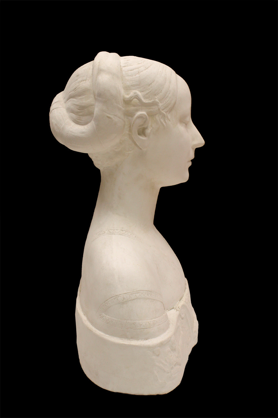 side view photo of white plaster cast sculpture of female bust cropped at shoulders with decorative base against black background